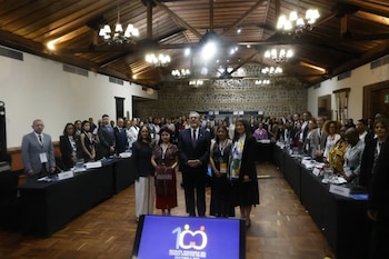 Un grupo de asistentes posa de pie alrededor de mesas en forma de U dentro de un salón con techo de madera y una pantalla frontal