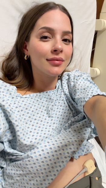 Mariana Rodríguez recostada en una cama clínica con bata estampada y un sensor médico con cable colocado sobre el abdomen