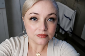 Primer plano de una mujer con cabello rubio, ojos azules, pendientes dorados de aro y un collar con dije de corazón.