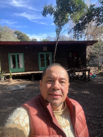 Hombre sonriente posa en primer plano frente a una casa de tierra y madera con techo de chapa y ventanas verdes, rodeada de árboles bajo cielo azul