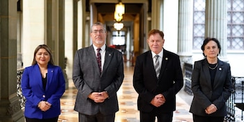 Cuatro personas vestidas con trajes formales están de pie en un pasillo con columnas de piedra, piso de mosaico y lámparas colgantes