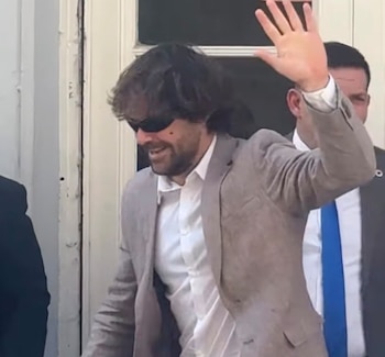 Un hombre con saco beige, camisa blanca y gafas oscuras levanta la mano izquierda en saludo frente a una puerta blanca