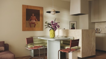 Comedor y cocina de diseño moderno. Mesa azul claro con jarrón amarillo y flores moradas, sillas rojas, banco verde. Un cuadro grande cuelga en la pared.