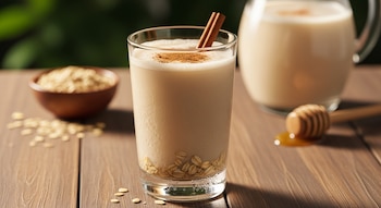 Un vaso de agua de avena con canela y un palo de canela, un tazón de avena, una jarra con la bebida y un cucharón de miel sobre una mesa de madera.