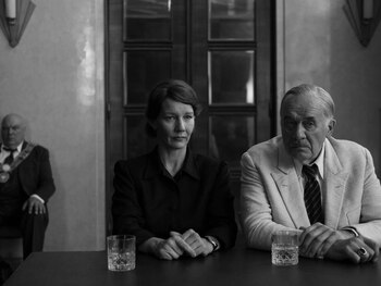 Imagen en blanco y negro: Una mujer y un hombre mayor sentados en una mesa, con vasos. Otro hombre se sienta en el fondo a la izquierda