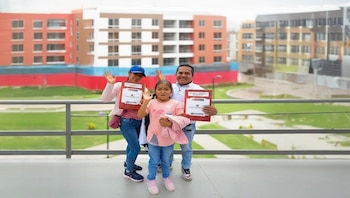 Hombre, mujer y niña sonrientes, con certificados del Gobierno del Perú y COFOPRI, frente a edificios de varios pisos y un área verde con camino