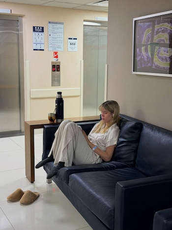 Mujer rubia sentada en sofá negro de cuero con pulsera de hospital. Termo y mate en mesa de madera. Ascensores y carteles de información