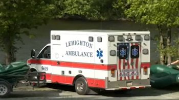 Ambulancia blanca con franjas rojas y logo 'Lehighton Ambulance', con el símbolo Estrella de la Vida y bandera estadounidense en puertas traseras, con un remolque y árboles
