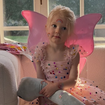 Niña pequeña rubia con un vestido de hada rosa con puntos brillantes, alas rosas, y pegatinas de arcoíris en la cara y brazos, sosteniendo un peluche gris