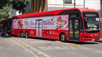 Un Metrobús rojo articulado con la leyenda '¡Feliz Día de las Madres!' y decoraciones florales. Se encuentra en una calle con la marca 'SOLO BUS' y personas al fondo.