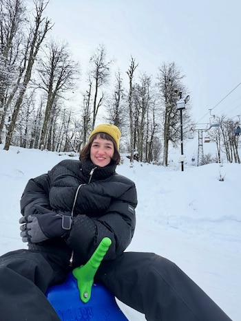 Una mujer sonriente con chaqueta negra, gorro amarillo y guantes grises está sentada en un trineo azul en un paisaje nevado con árboles y un telesilla