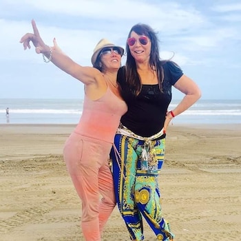 Dos mujeres posan en una playa de arena frente al mar, una lleva sombrero y la otra usa anteojos de sol