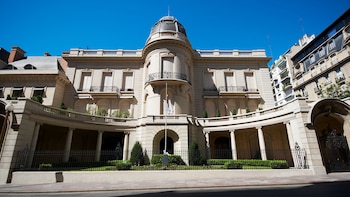 Edificio de estilo clásico con una cúpula central, balcón, reja frontal, pilares y una bandera en un mástil frente a un cielo despejado
