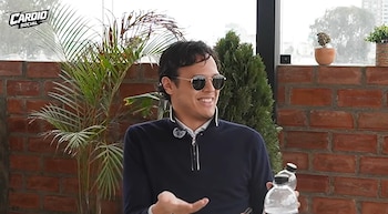 Hombre con gafas de sol y suéter azul marino sentado ante una pared de ladrillo con plantas de fondo