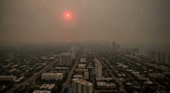 Vista aérea de ciudad con edificios, carreteras, vegetación, océano y un sol rojizo brillante, todo cubierto por una capa de humo denso.