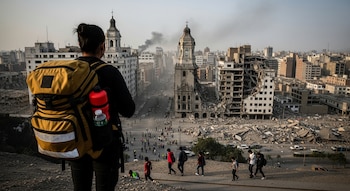 Vista trasera de una persona con mochila amarilla y botella roja, mirando una ciudad con edificios destruidos y en pie, humo en el horizonte, y personas caminando.