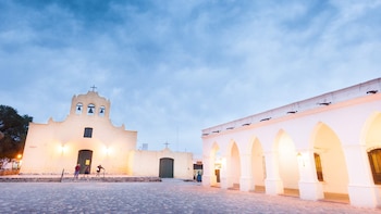 Una iglesia colonial con campanario y un edificio de arcos blancos iluminados al atardecer. Unas personas están en la plaza de adoquines frente a la iglesia. Cachi, Salta pueblo nominado para los Best Tourism