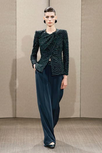 Una mujer desfila en una pasarela con chaqueta verde texturizada, pantalones anchos azul oscuro y zapatos planos verdes. Lleva aretes negros y el cabello recogido. El fondo es liso beige