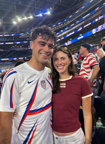 Primer plano de Gio Reyna sonriente con una camiseta de fútbol de EE. UU. y su esposa sonriente con una camiseta granate, ambos en un estadio