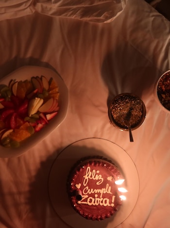 Un pastel de chocolate con velas encendidas, un plato de frutas cortadas y un mate sobre una cama con sábanas blancas