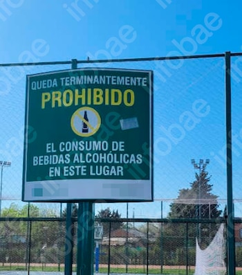 Cartel verde con texto 'PROHIBIDO EL CONSUMO DE BEBIDAS ALCOHÓLICAS' y un icono de botella tachada, montado en una cerca de malla metálica