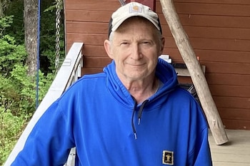 Hombre de edad mediana con gorra clara y sudadera azul con pin en el pecho posa sonriendo. Detrás, baranda blanca, pared marrón y vegetación verde