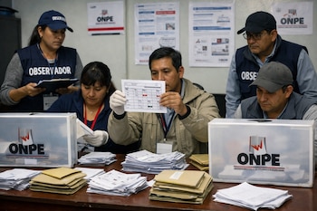 Personas con chalecos de 'OBSERVADOR' y guantes revisan boletas de votación y sobres en una mesa con urnas transparentes de la ONPE. Al fondo, afiches informativos.