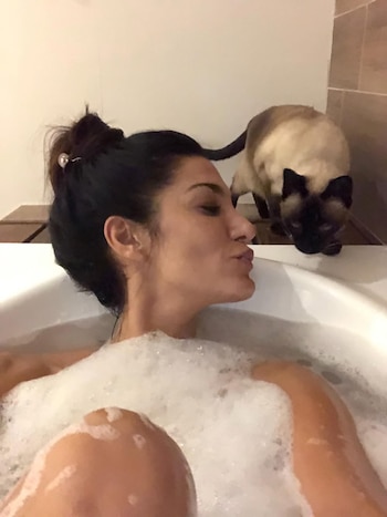 Silvina Escudero en una bañera llena de espuma, con los labios fruncidos, mientras su gata siamesa Afrika la observa desde el borde