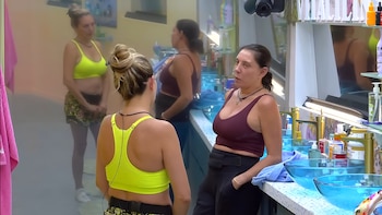 Mariana Ochoa, con top deportivo amarillo, habla con Cynthia Klitbo, vestida con top oscuro, en un baño con lavabos azules