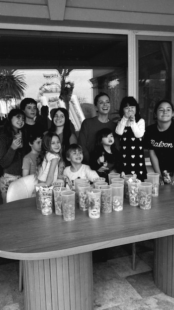 Imagen en blanco y negro de una mujer sonriente con varios niños, algunos sonriendo y sosteniendo vasos con dulces, detrás de una mesa con muchos vasos transparentes