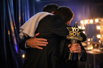 Dos personas se abrazan con la espalda hacia la cámara; uno lleva una toalla blanca en el hombro y el otro sostiene un trofeo dorado y negro de "Yo soy grandes batallas".