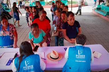 Una fila de personas espera ante una mesa donde dos voluntarios con chalecos azules las atienden