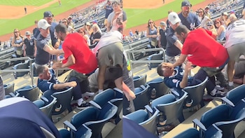 Una pelea en las gradas de un estadio de béisbol con varios hombres involucrados, uno de camiseta roja golpeando a otro sentado de camiseta azul