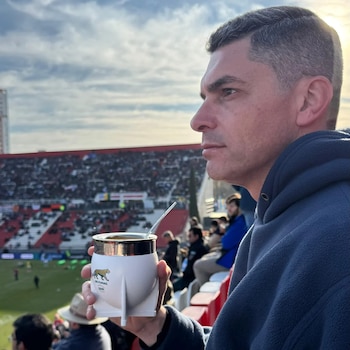 Hombre de perfil sosteniendo un mate blanco con bombilla metálica en la tribuna de un estadio con público en el fondo