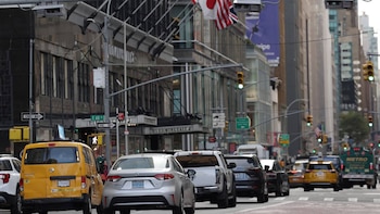 Vista trasera de varios vehículos, incluyendo taxis amarillos y autos plateados, esperando en el tráfico con edificios urbanos y semáforos verdes al fondo
