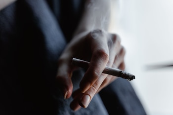 Mano de persona adulta con un cigarrillo encendido que tiene ceniza en la punta y emite humo blanco. Fondo claro y tela oscura