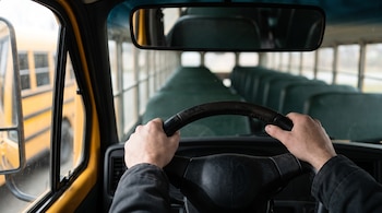Manos sobre el volante negro de un autobús escolar amarillo con asientos verdes vacíos al fondo.
