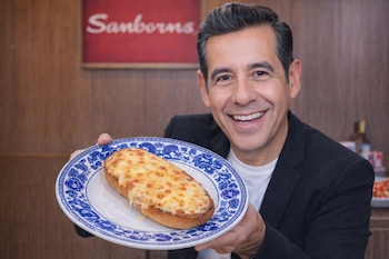 Yordi Rosado, un hombre de piel morena clara y cabello oscuro peinado hacia atrás, sonríe a cámara sosteniendo un plato de talavera azul con un mollete de queso derretido.