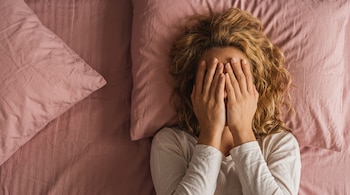 Una mujer de cabello rubio permanece acostada en una cama con sábanas rosas mientras se cubre la cara con ambas manos.
