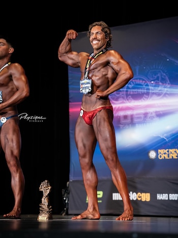 Joseph Baena posa en un escenario de fisicoculturismo, sonriendo y flexionando el bíceps. Lleva bañador rojo, medalla y su piel está bronceada