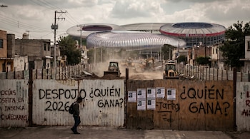 Un hombre camina frente a una valla con grafitis que dice "DESPOJO ¿QUIÉN GANA?". Detrás, maquinaria de construcción y tres estadios modernos bajo un cielo nublado.