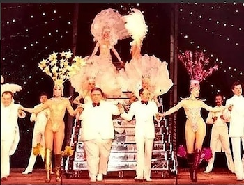 Escenario con luces, Alberto Olmedo, Jorge Porcel, Moria Casán y Susana Giménez con vestuario de revista, otros bailarines y un fondo oscuro