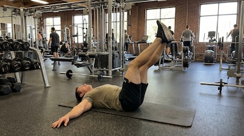 Un hombre acostado boca arriba en una colchoneta gris realiza elevaciones de piernas en un gimnasio, con otros usuarios y máquinas de ejercicio al fondo.