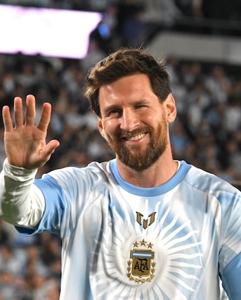 Lionel Messi sonríe con la mano derecha levantada y viste una camiseta celeste y blanca con el escudo de la AFA