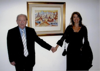 Hombre mayor y mujer adulta tomados de la mano posan de pie frente a una pared blanca. Detrás, un cuadro enmarcado con una escena de montañas y edificios