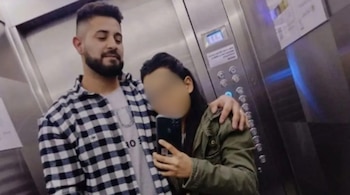 Un hombre con barba y camisa a cuadros abraza a una mujer de cabello oscuro con el rostro pixelado dentro de un ascensor con paredes metálicas