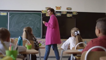 Uma professora de vestido rosa e jeans em frente a um quadro negro e uma tela preta, com uma aluna sentada em uma mesa na frente.