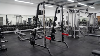 Interior de un gimnasio con una máquina Smith, estantes de mancuernas, bancos de ejercicio y máquinas de poleas sobre un suelo de caucho negro.
