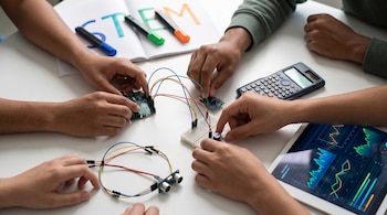Manos diversas manipulan componentes de robótica, sensores y cables sobre una mesa blanca. Una calculadora y una tableta con gráficos encendida. Cuaderno con 'STEM' desenfocado.