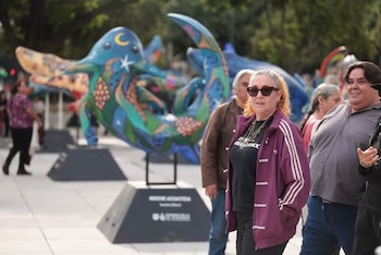 Una mujer con gafas de sol y chaqueta deportiva, personas y esculturas de criaturas marinas con diseños coloridos en pedestales oscuros en un espacio público